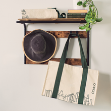 Load image into Gallery viewer, Tote bag with city skyline design hanging on a wooden rack with a plant and books in the background.
